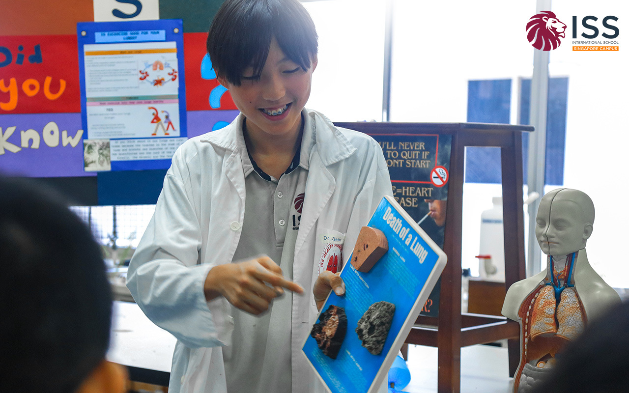 An MYP student in a lab coat presents a "Death of a Lung" poster in a classroom setting